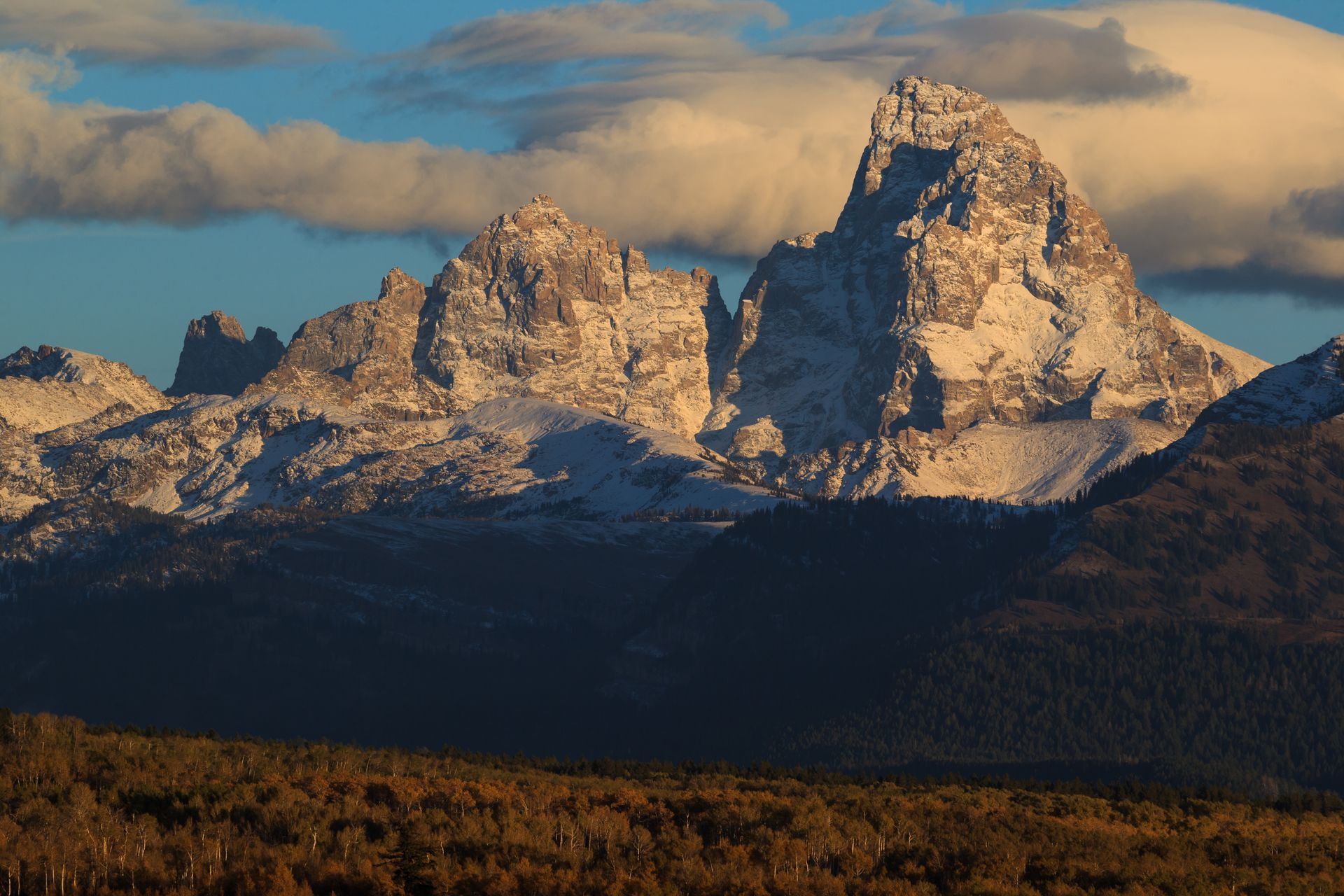 Dramatic rocky mountain peaks with snow and forested foothills under blue sky with clouds