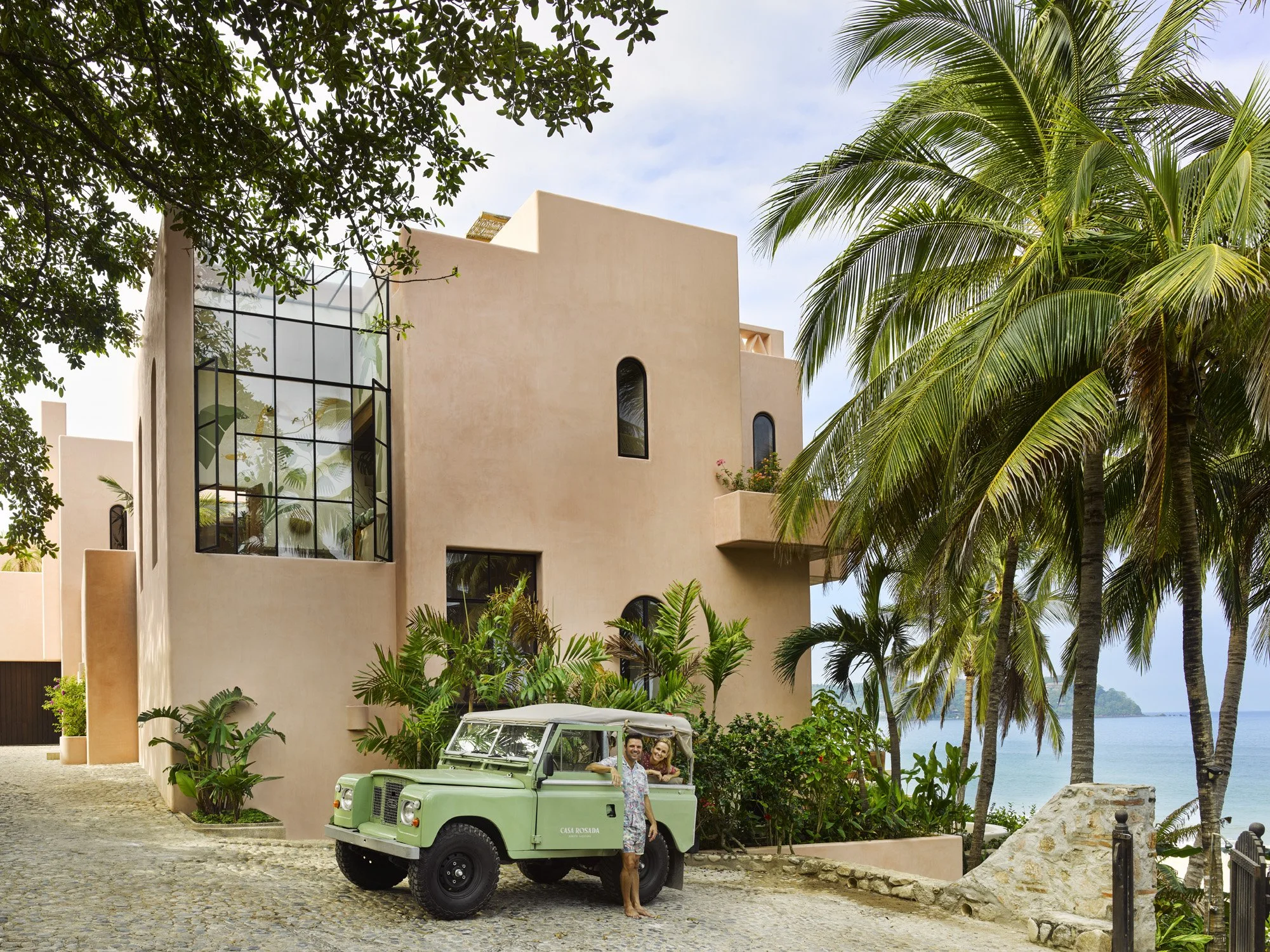 Beige modern villa with palm trees and turquoise ocean view, green vintage Land Rover parked in front