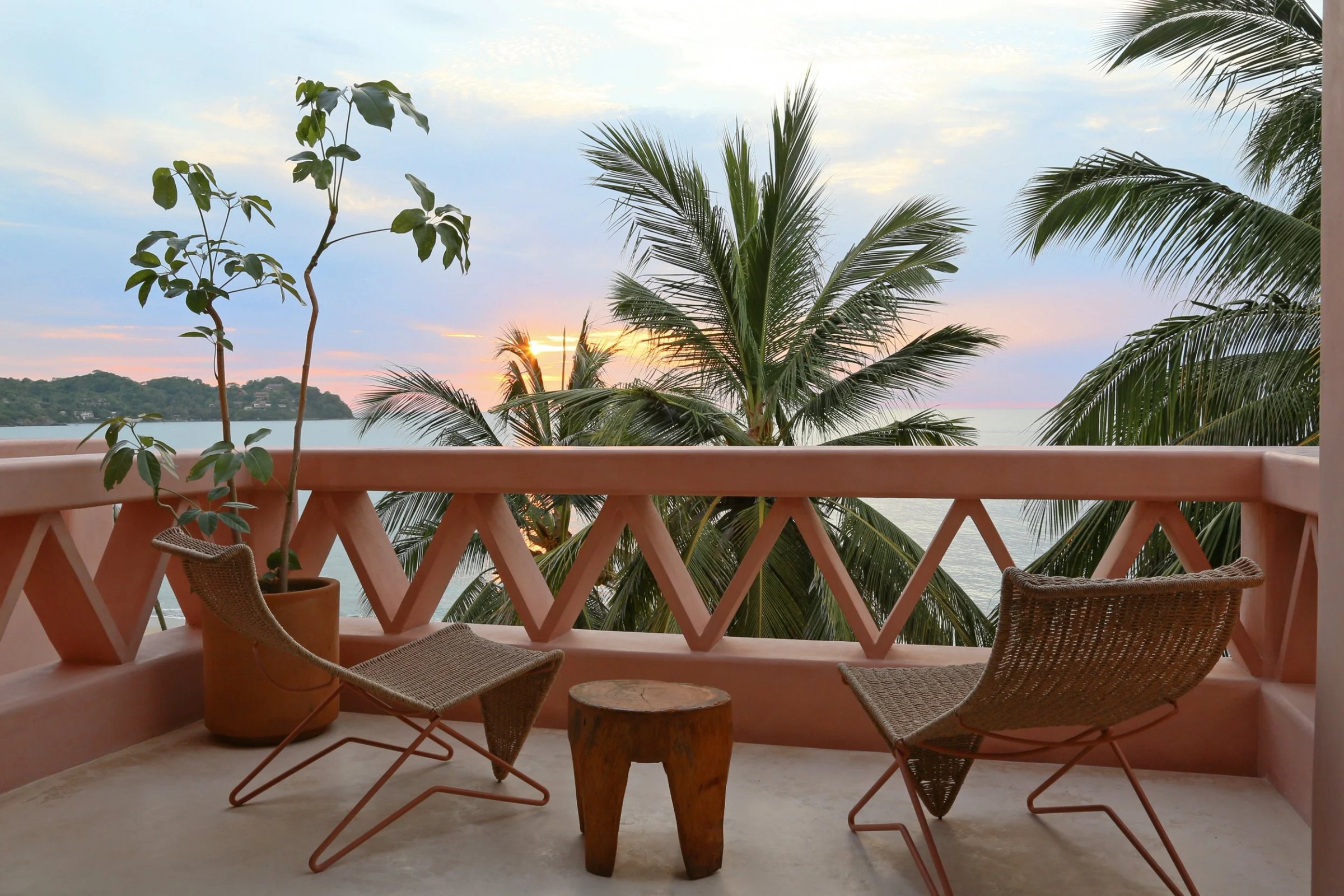 Terracotta balcony with woven chairs overlooking tropical bay at sunset, palm trees and potted plants visible