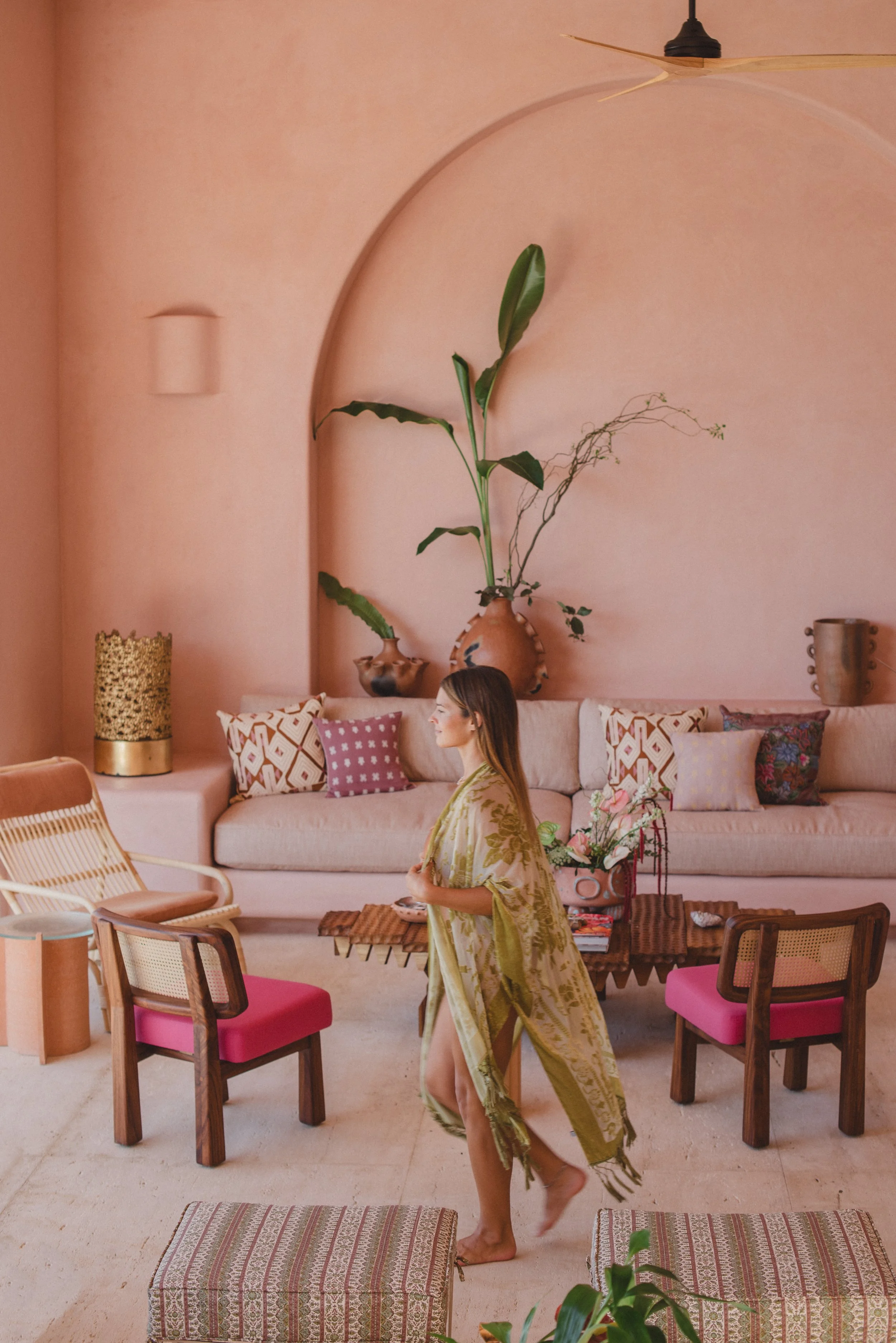 Pink living room with arched alcove, pink sofa, wooden chairs with pink cushions, potted plants, and woman in yellow robe