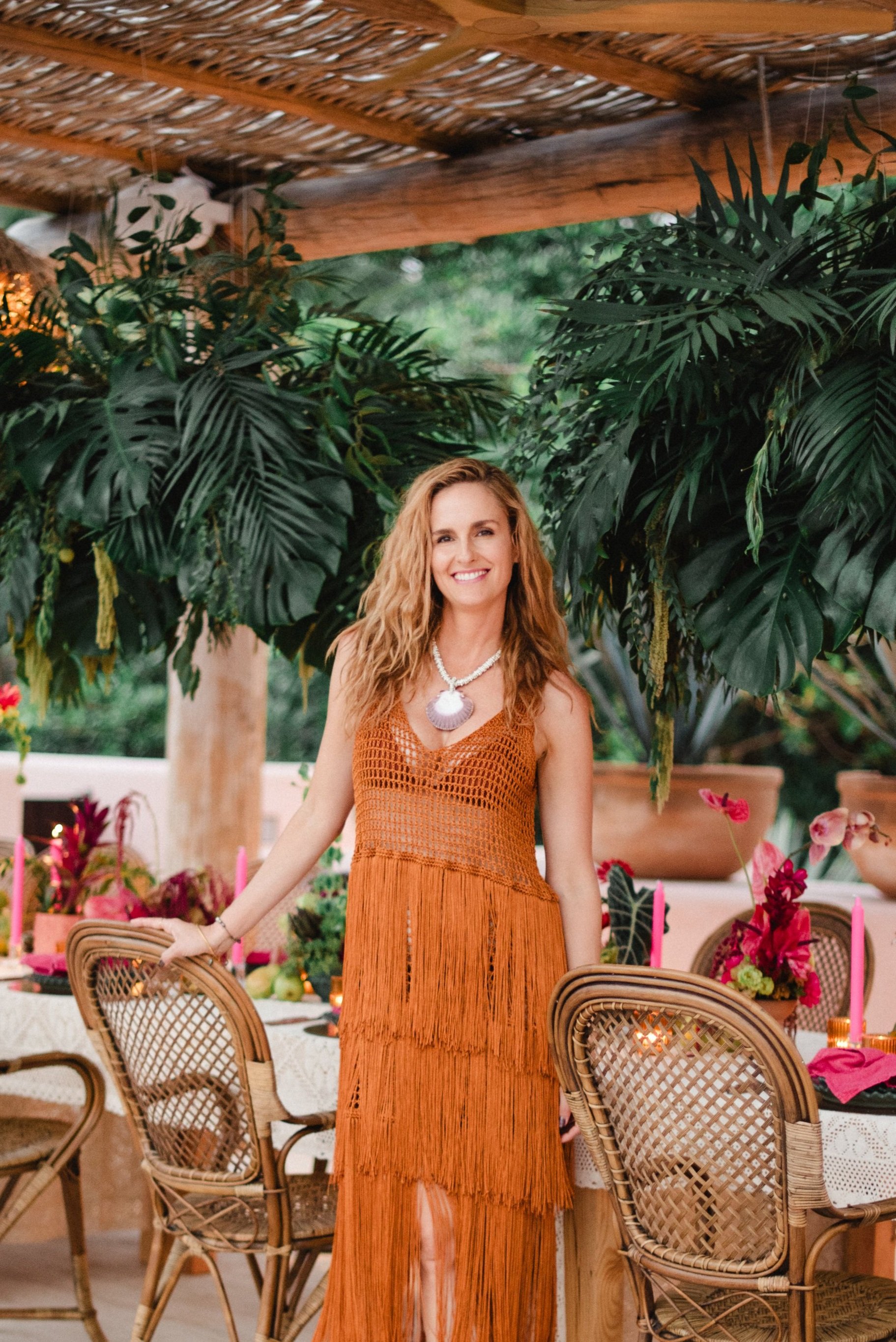 Woman in orange fringe dress standing in tropical garden patio with rattan chairs and hanging plants