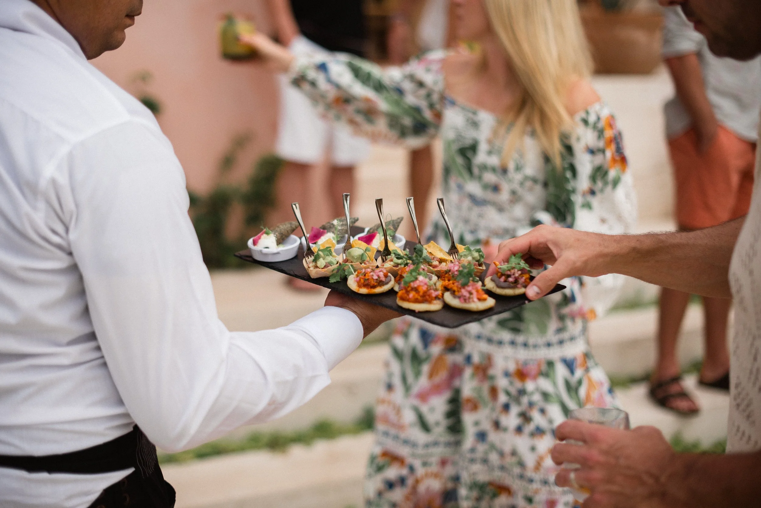 Server in white shirt holding black tray of appetizers with forks at outdoor gathering with guests