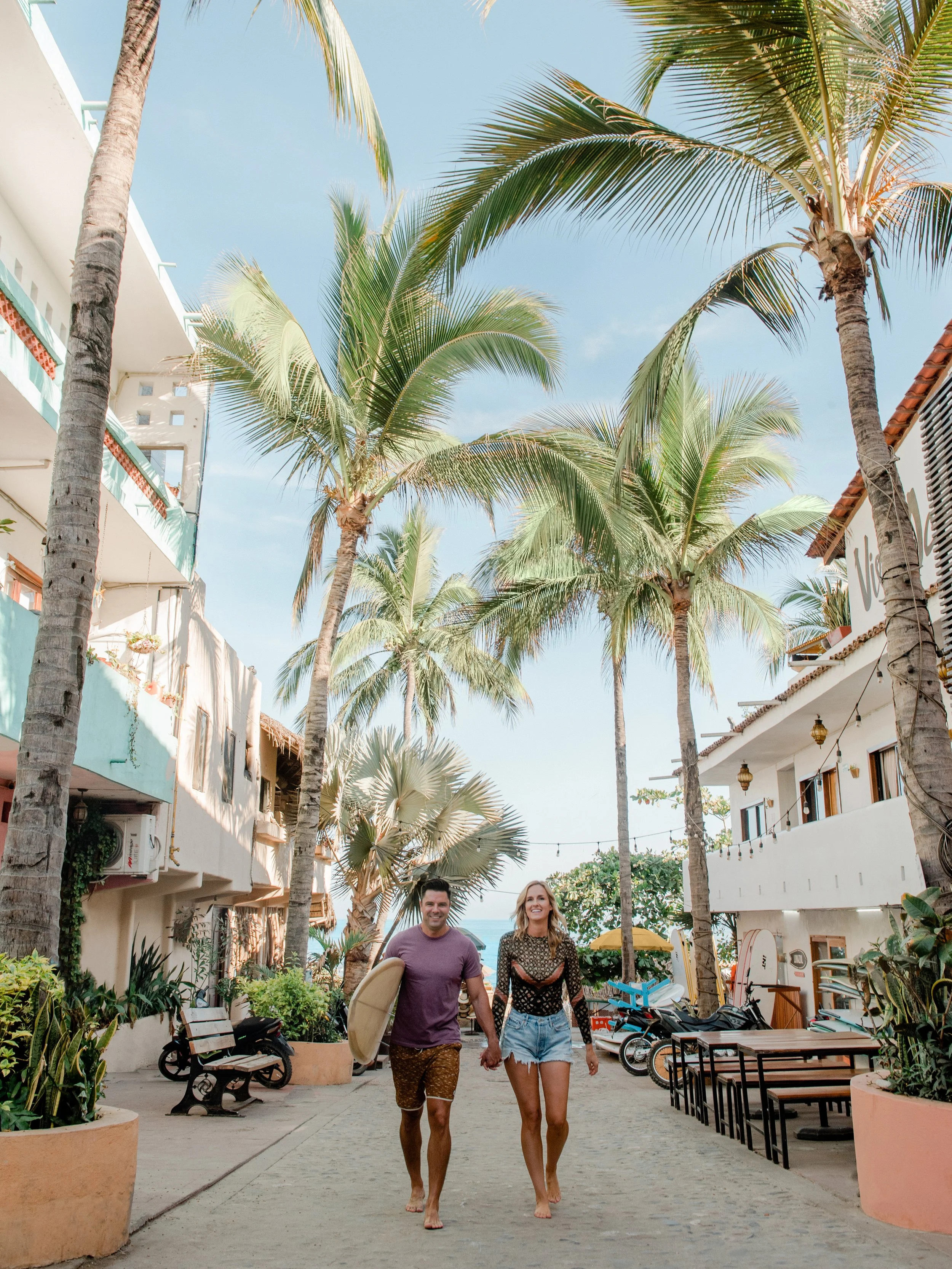 Couple walking barefoot through palm-lined courtyard with white buildings and clear blue sky