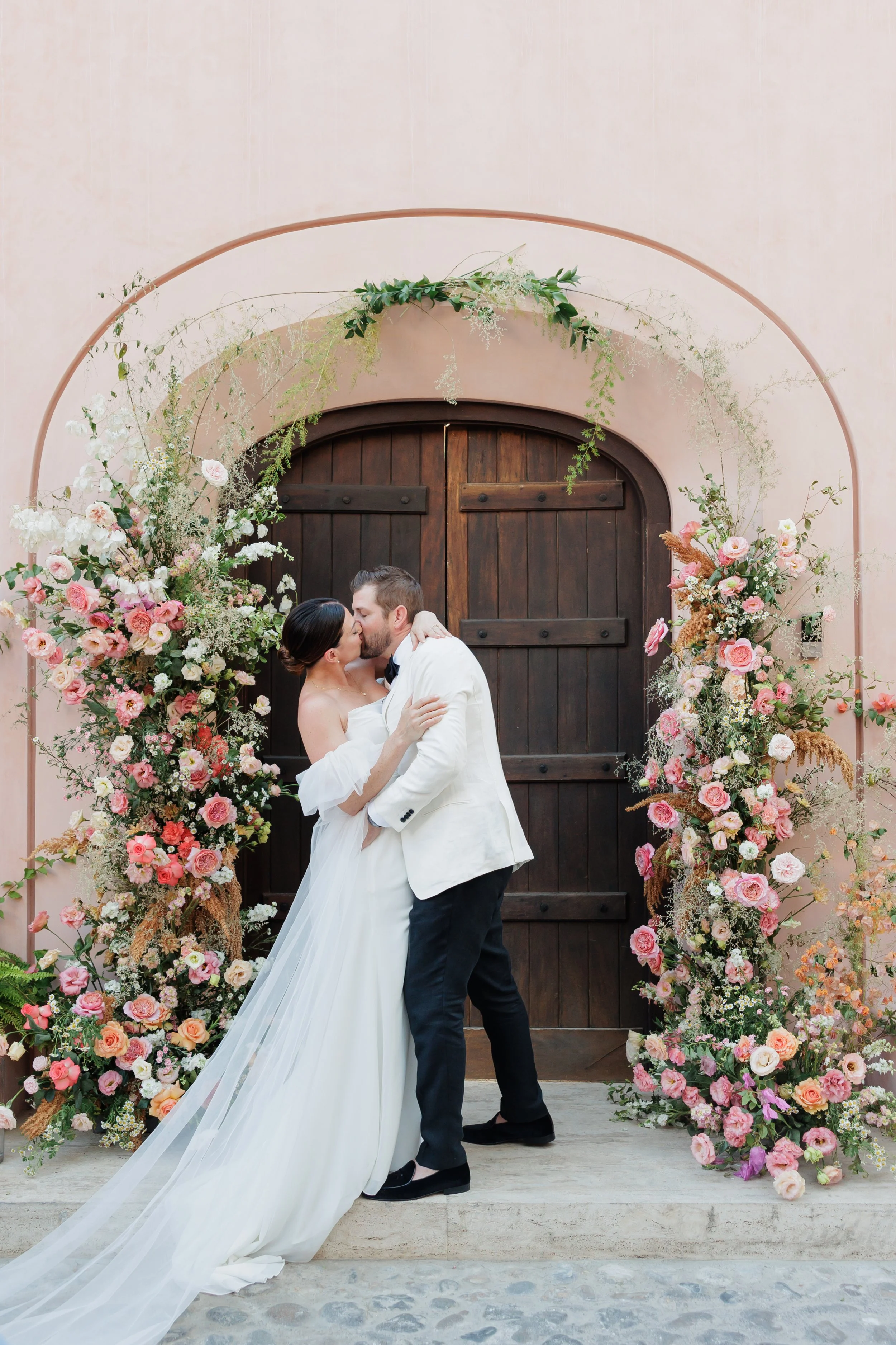 Bride and groom kissing under arched doorway decorated with pink and white flowers