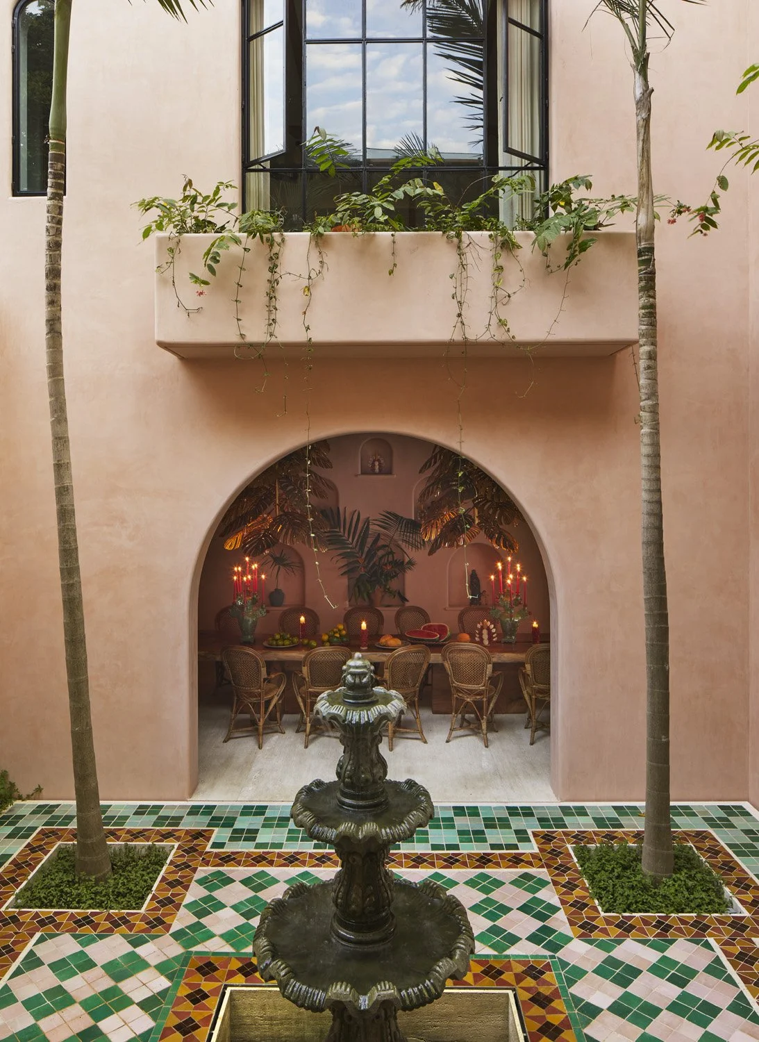Courtyard with pink stucco walls, arched alcove containing dining table with candles, ornamental fountain, geometric tile floor, and potted plants on window