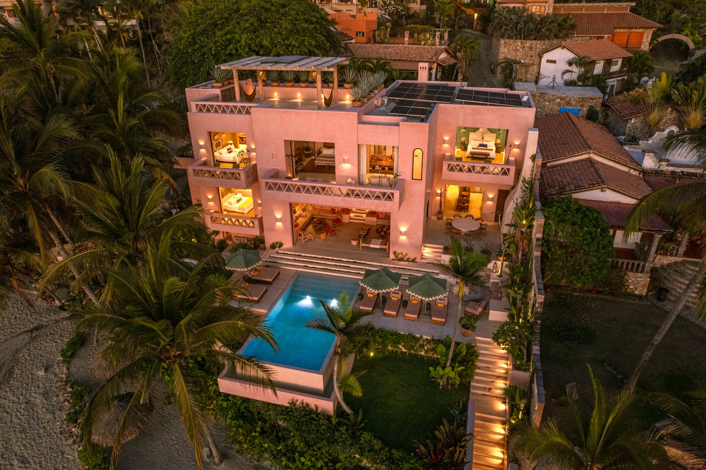 Aerial view of pink modern villa with illuminated pool, lounge chairs, and solar panels at dusk