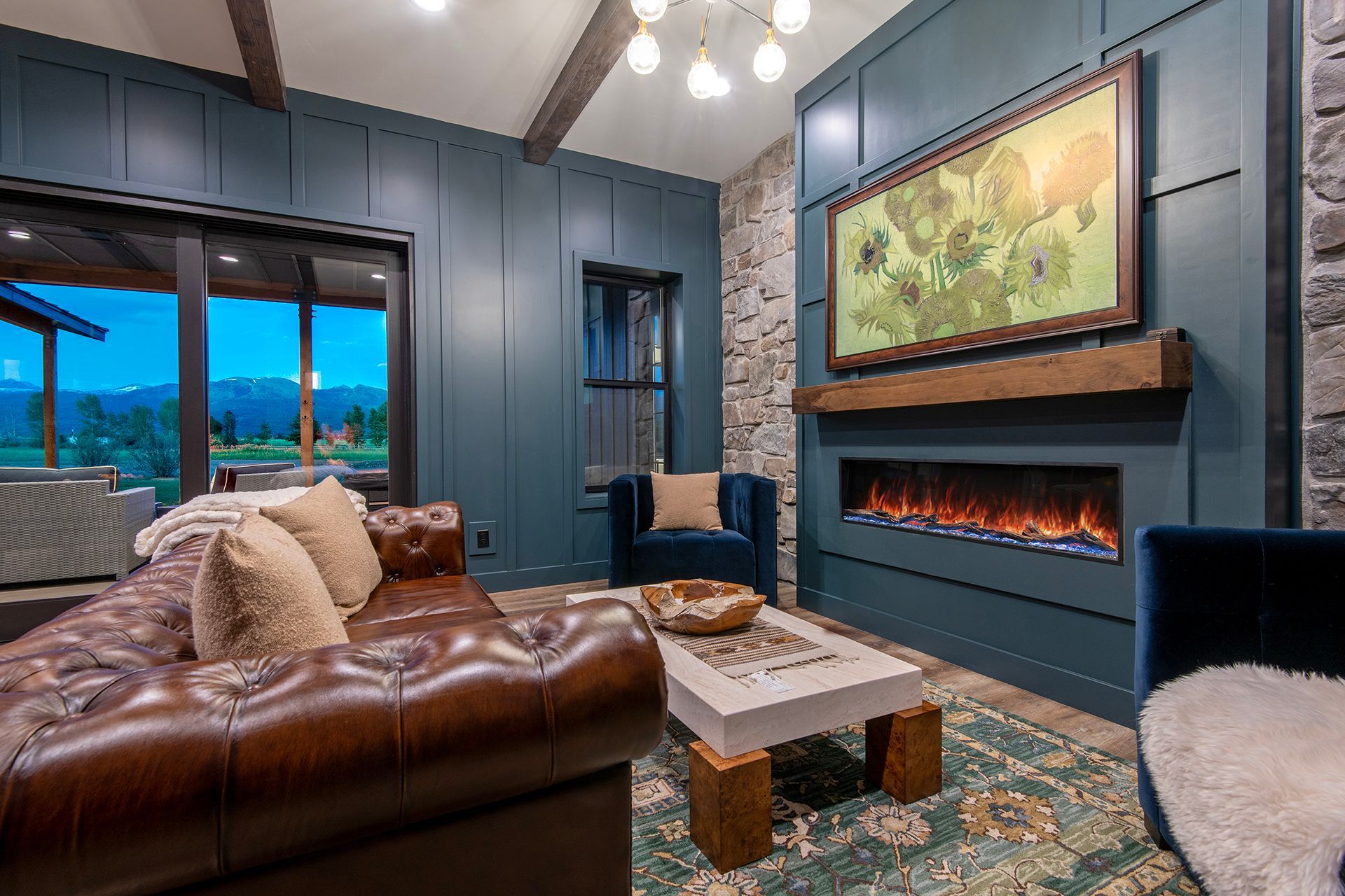 Modern living room with brown leather sofa, electric fireplace, blue-green walls, stone accent wall, and mountain views through glass doors.