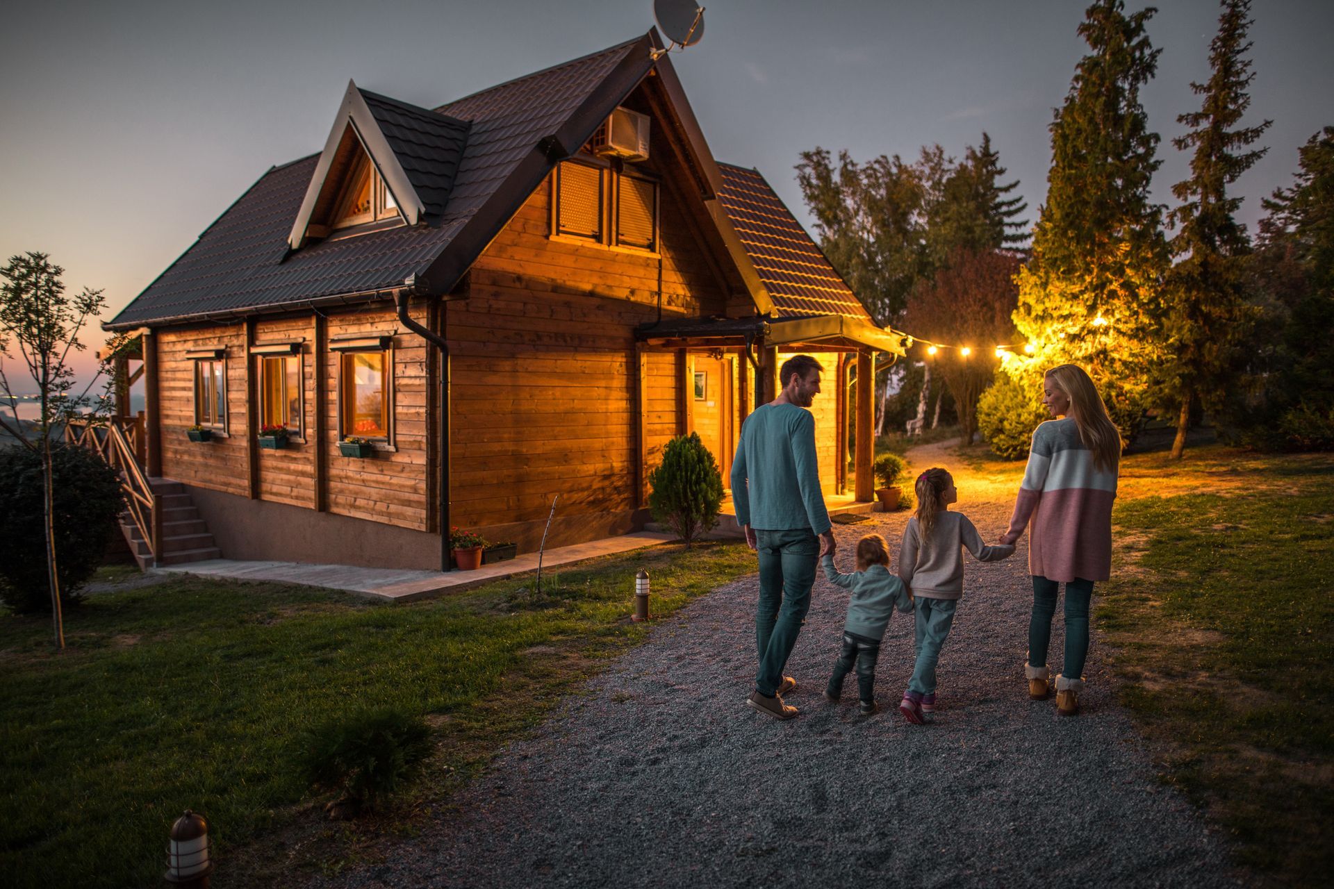 Illuminated wooden cabin at dusk with family of four standing on gravel path, surrounded by evergreen trees