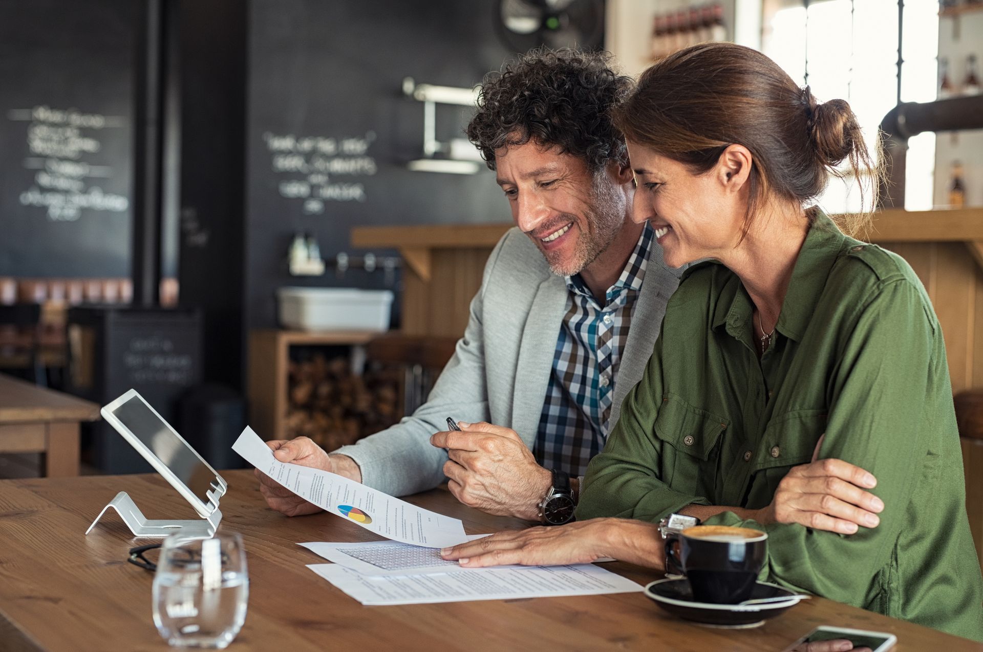 Two people reviewing documents and tablet at wooden table in modern café with black menu board backdrop