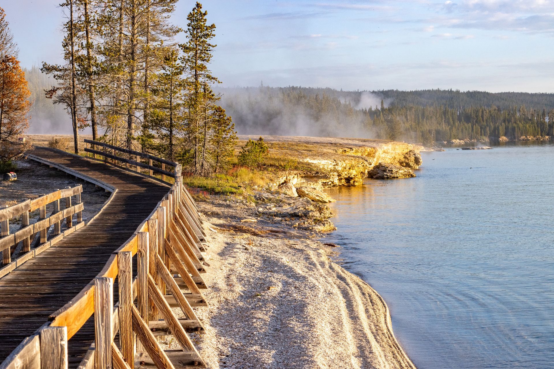 Wooden boardwalk along lake shore with geothermal springs steaming in distance, coniferous forest and rocky shoreline