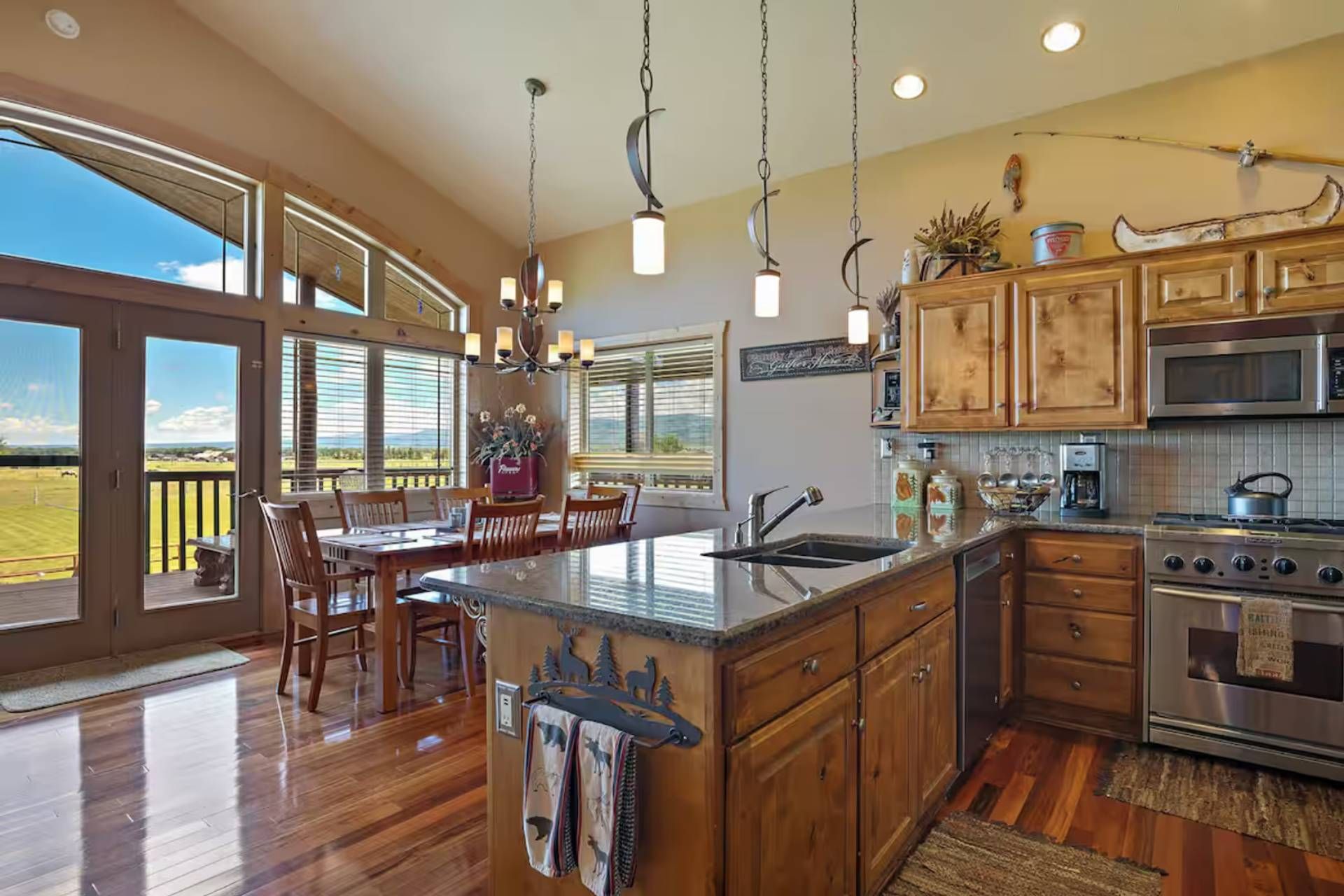 Open-plan kitchen and dining area with wood cabinetry, granite countertops, stainless steel appliances, and expansive windows overlooking green fields and