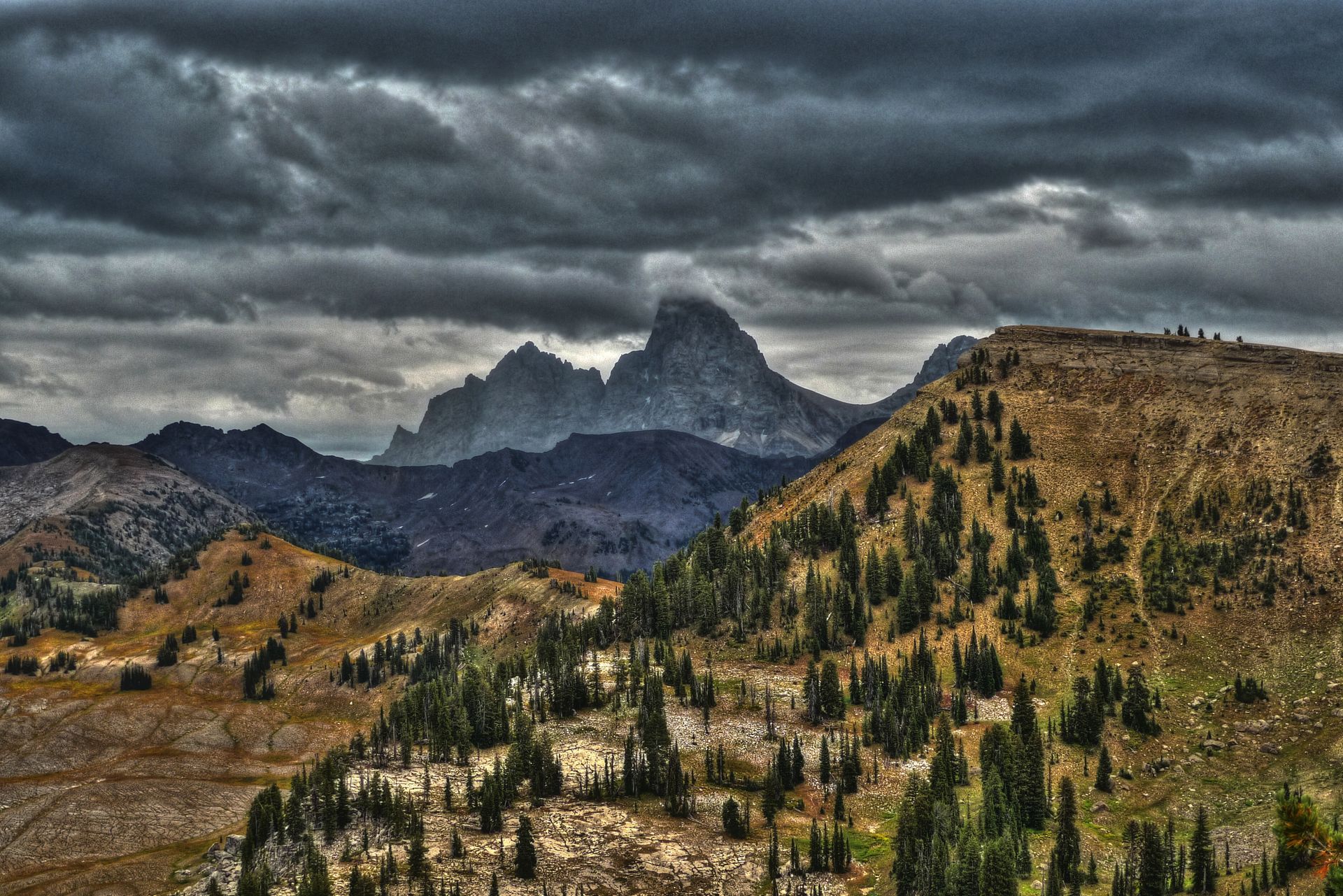 Alpine mountain landscape with jagged peaks, coniferous forest, and dramatic storm clouds overhead