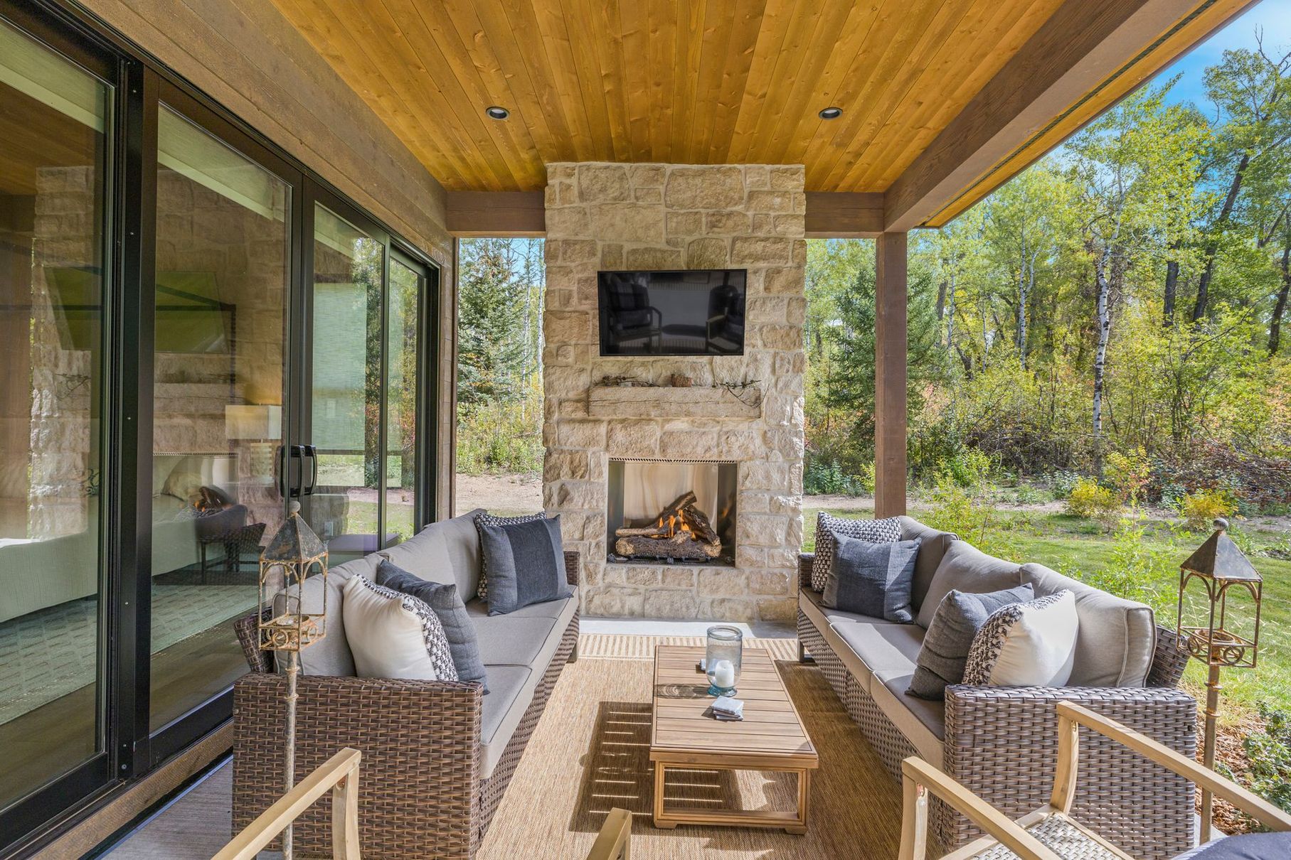 Covered patio with stone fireplace, TV, woven seating, wooden ceiling, and wooded views