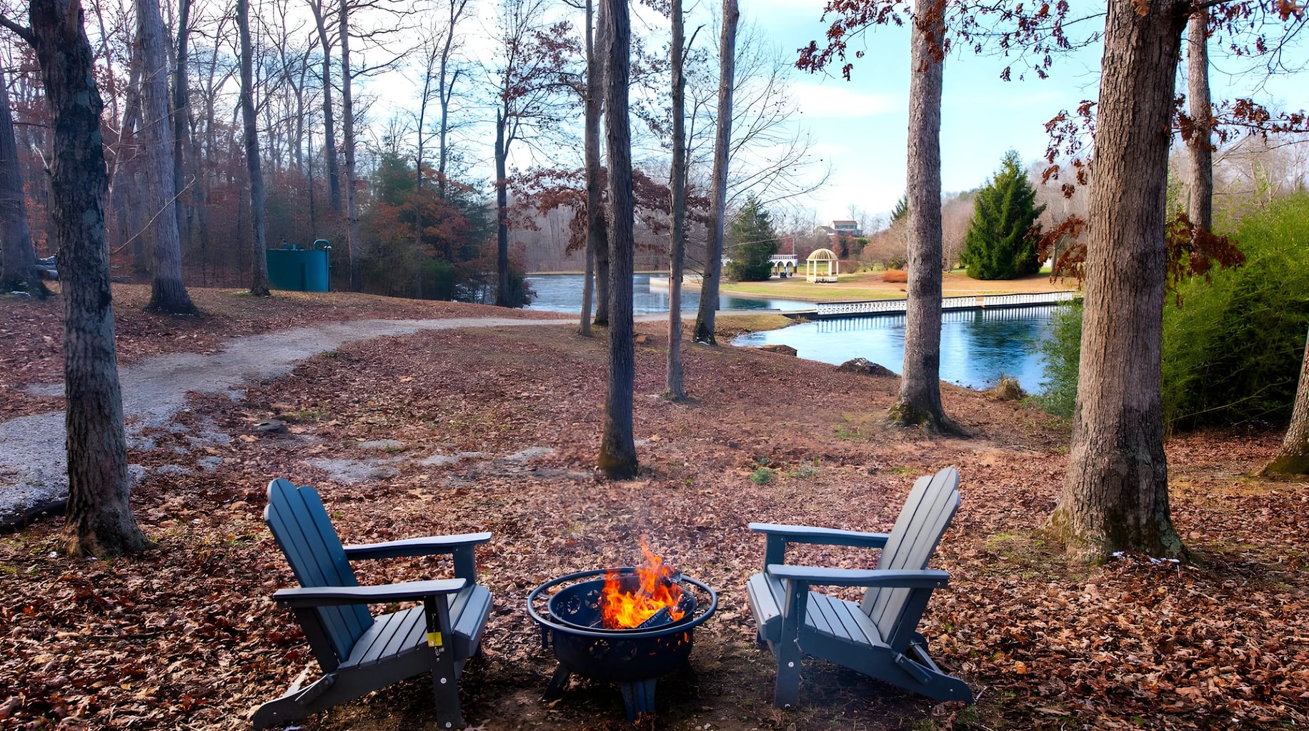 Two Adirondack chairs facing a fire pit with burning flames, overlooking a pond and wooded property with distant house.
