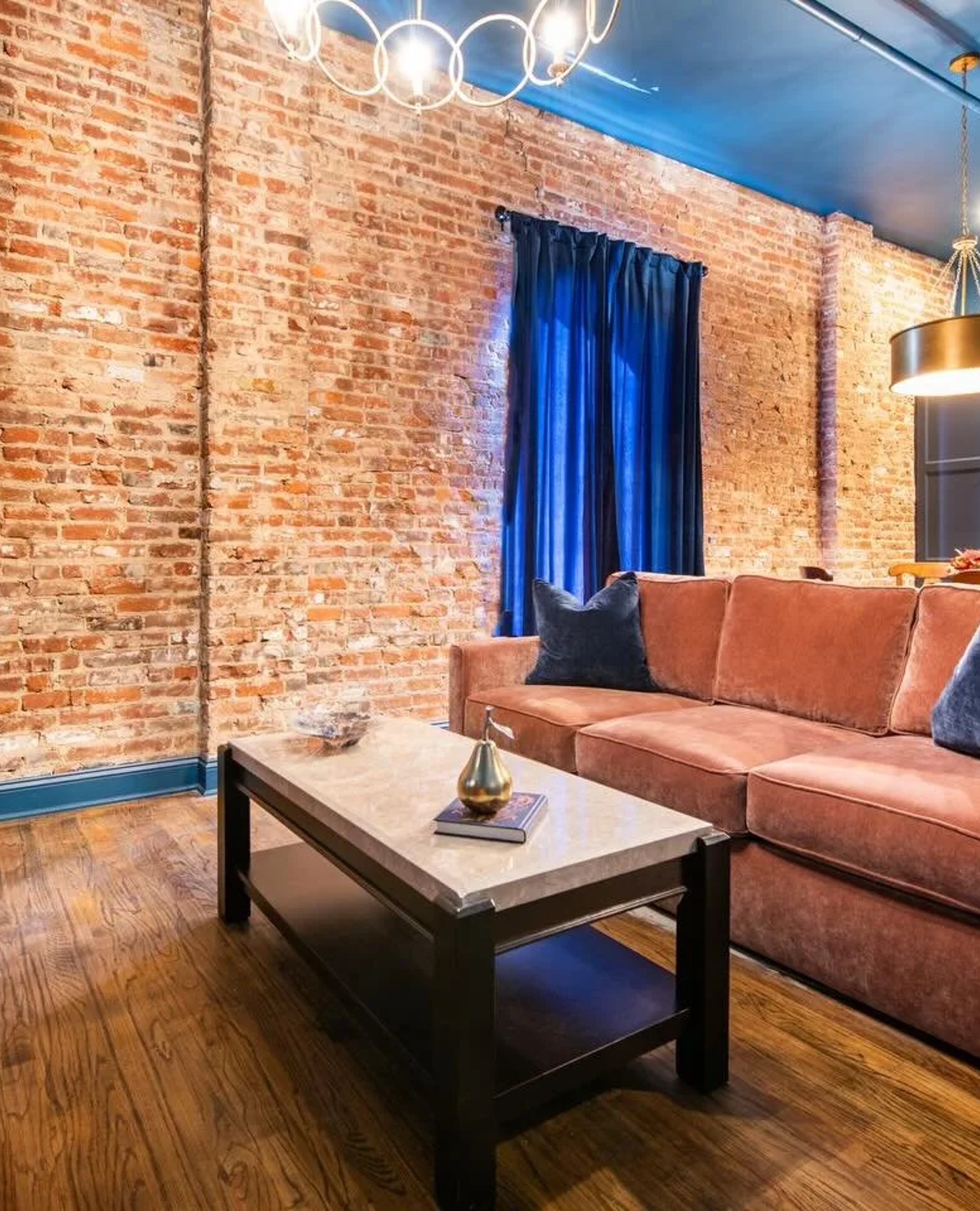 Modern loft living room with exposed brick walls, rust-colored sofa, marble coffee table, and blue curtains