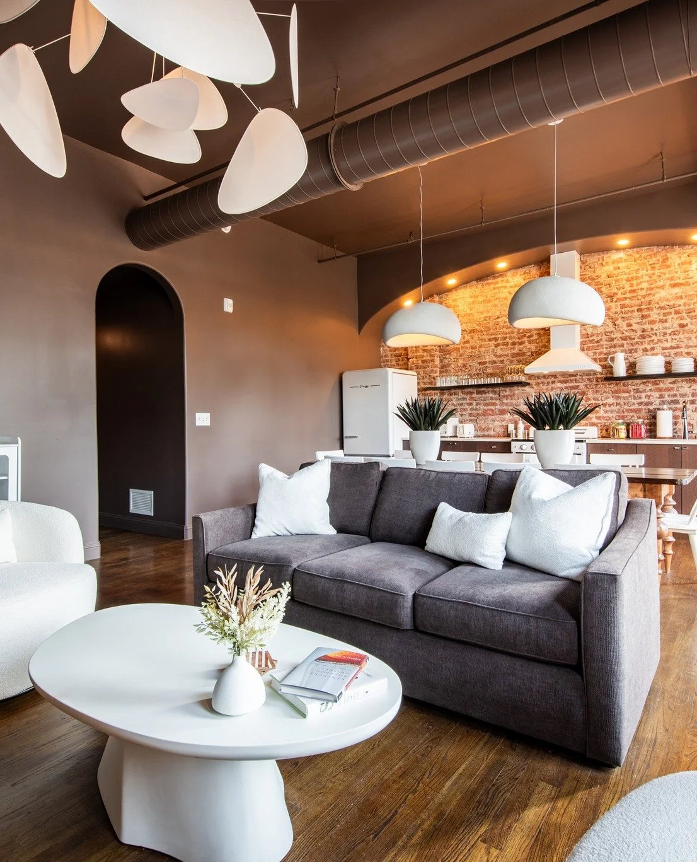 Modern loft living room with gray sofa, white round table, exposed brick wall, pendant lights, and industrial ceiling ductwork.