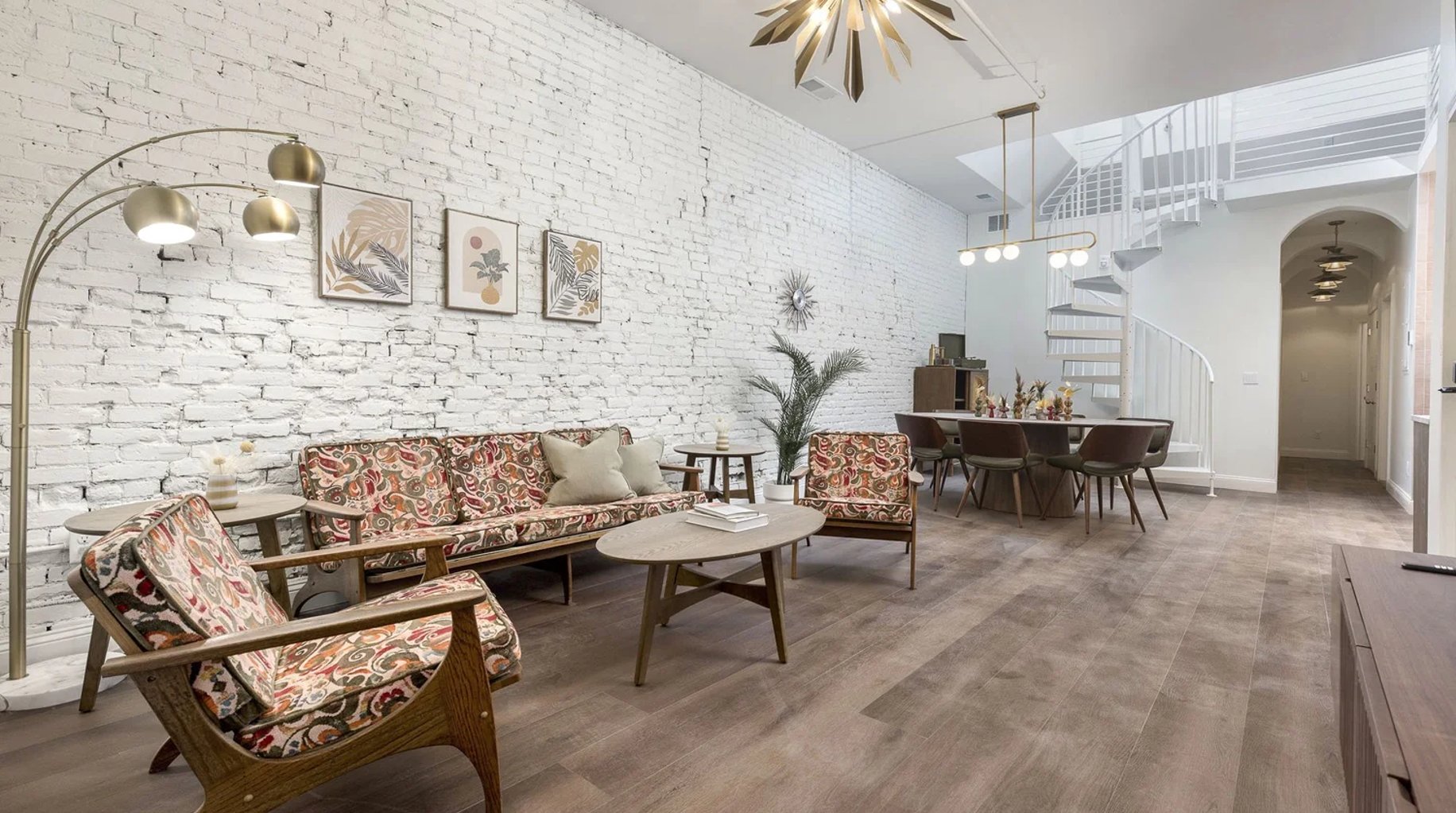 Modern loft living room with white brick walls, mid-century furniture in red patterned fabric, brass arc lamp, and spiral staircase visible in background.