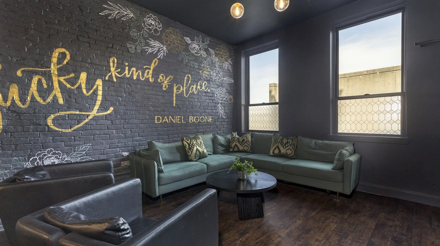 Modern lounge with dark brick wall featuring gold inspirational quote, gray and teal sectional sofa, pendant lights, and large windows overlooking landscape.