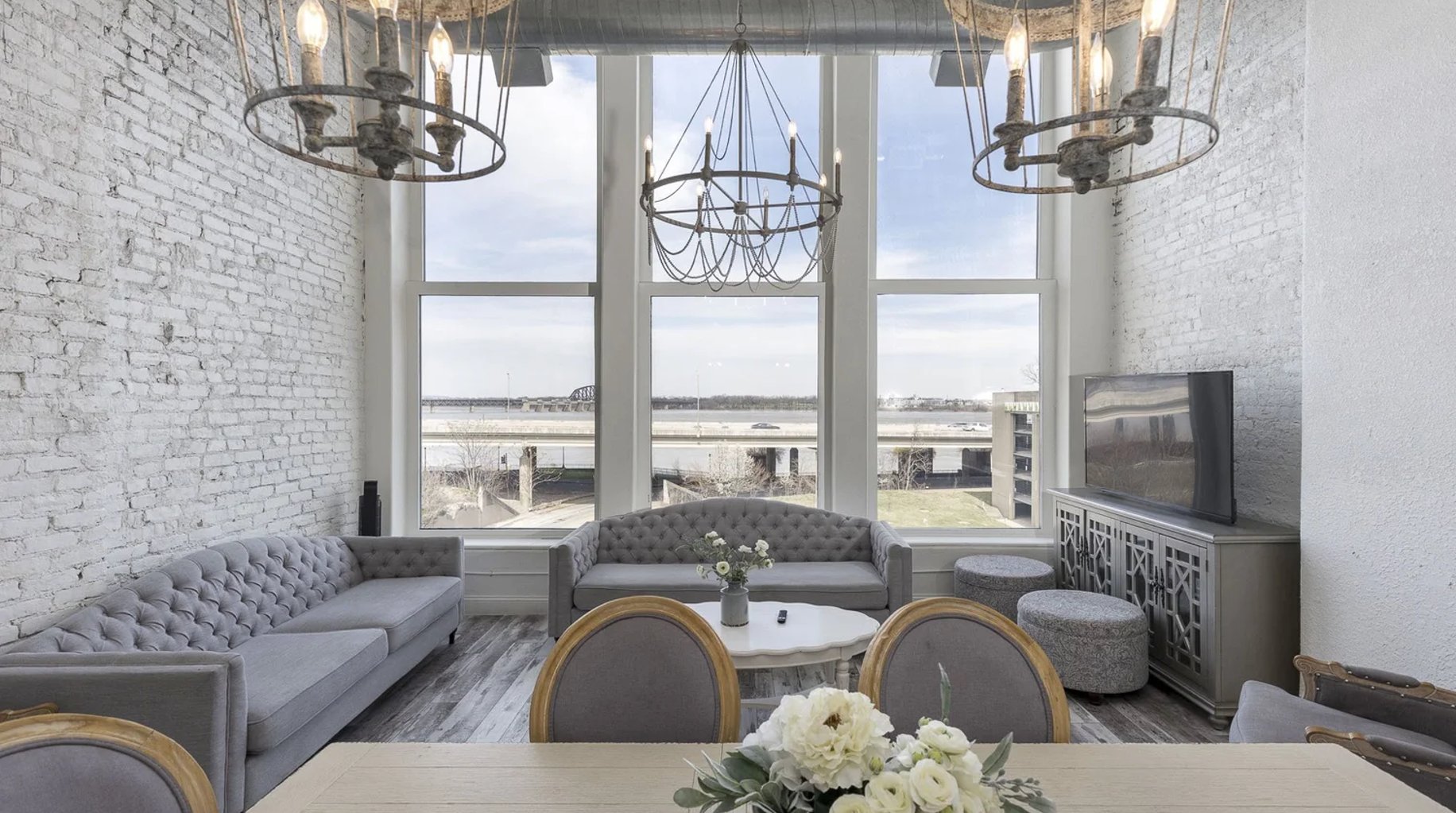 Modern living room with white brick walls, gray tufted seating, industrial chandeliers, and floor-to-ceiling windows overlooking a river.