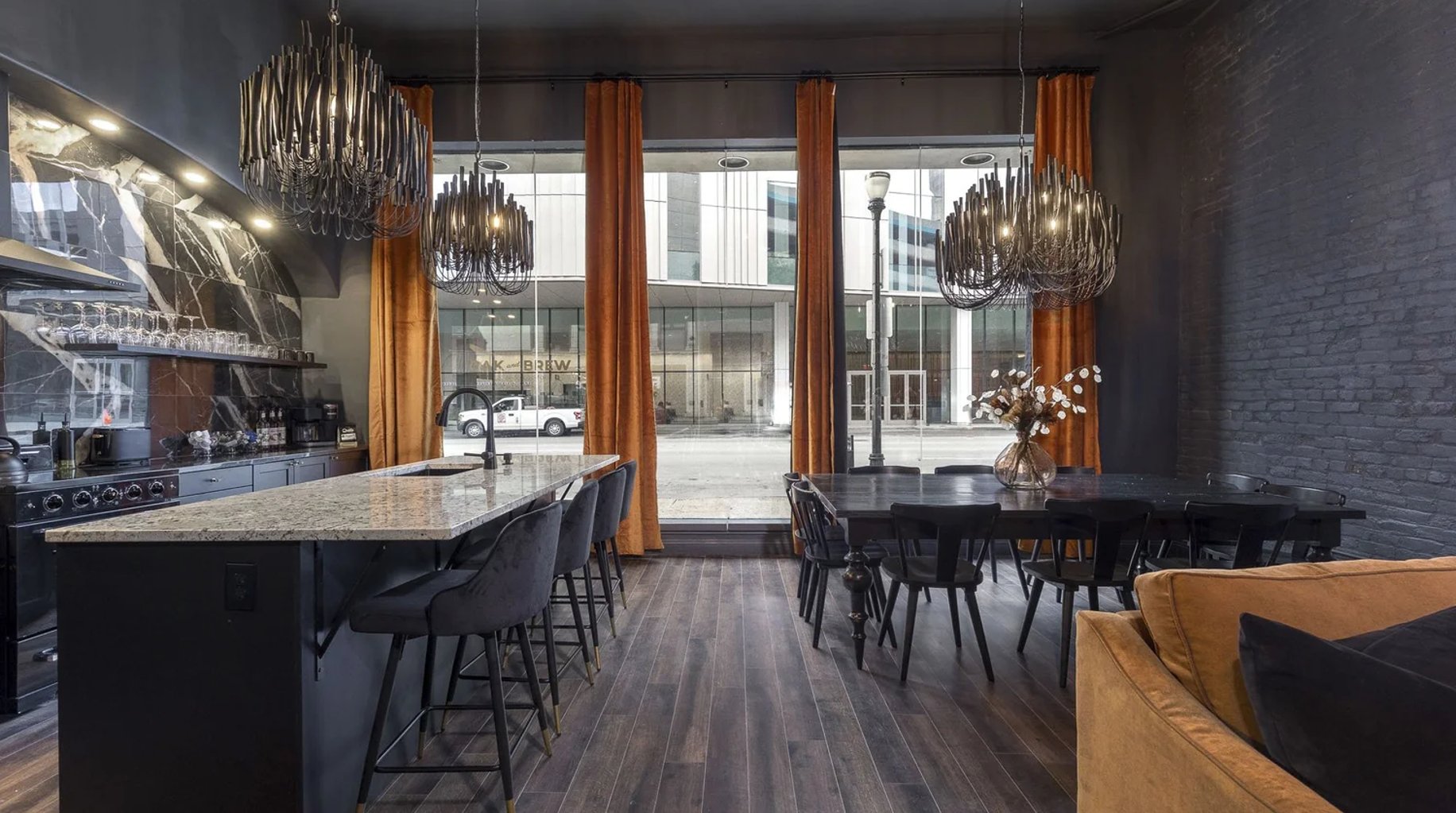 Modern open-plan kitchen and dining area with dark cabinetry, granite countertops, pendant chandeliers, and floor-to-ceiling windows overlooking urban street.
