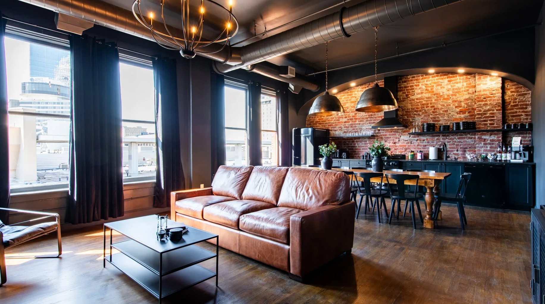 Industrial loft with cognac leather sofa, exposed brick kitchen, metal pendant lights, and city views through floor-to-ceiling windows.