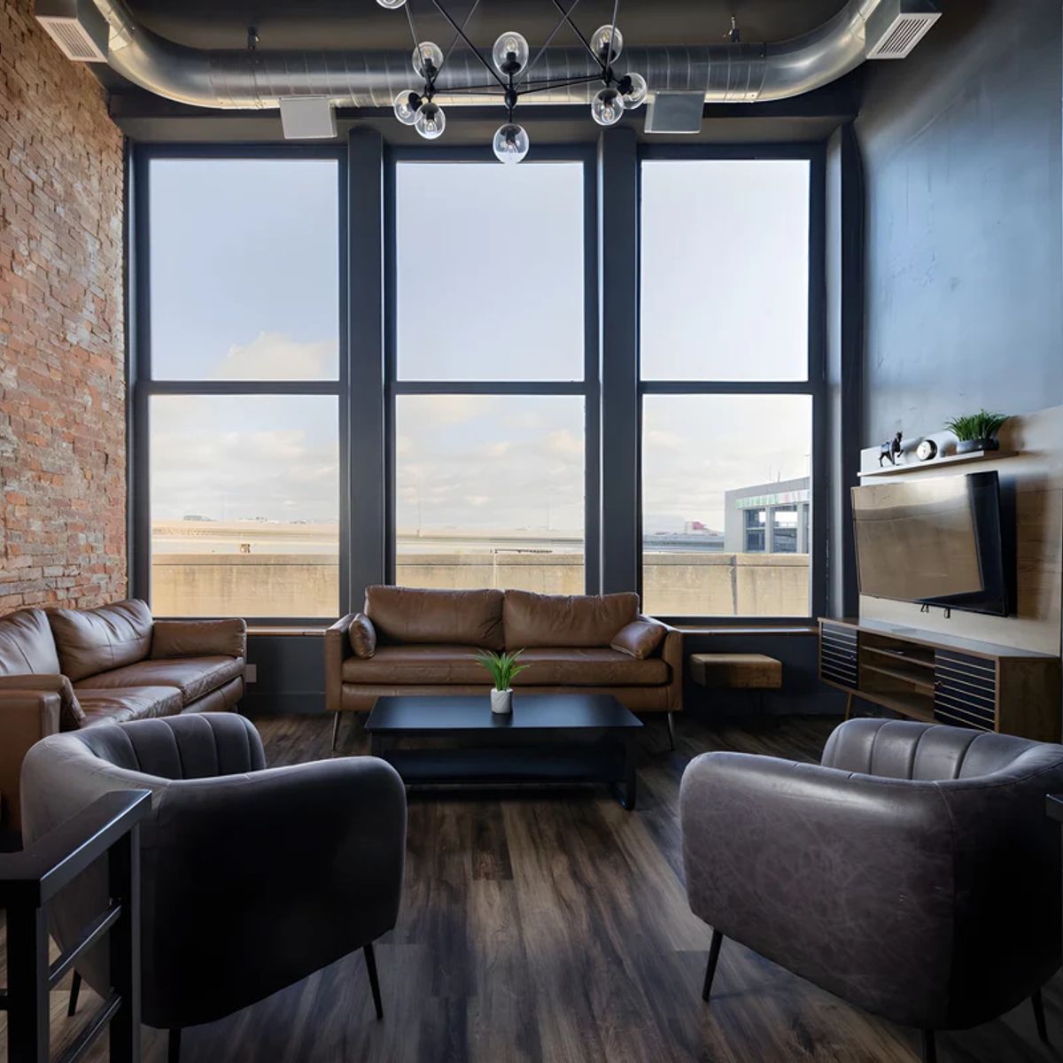 Modern loft living room with large windows overlooking waterfront, brown leather sofa, dark armchairs, and industrial ceiling with pendant lights